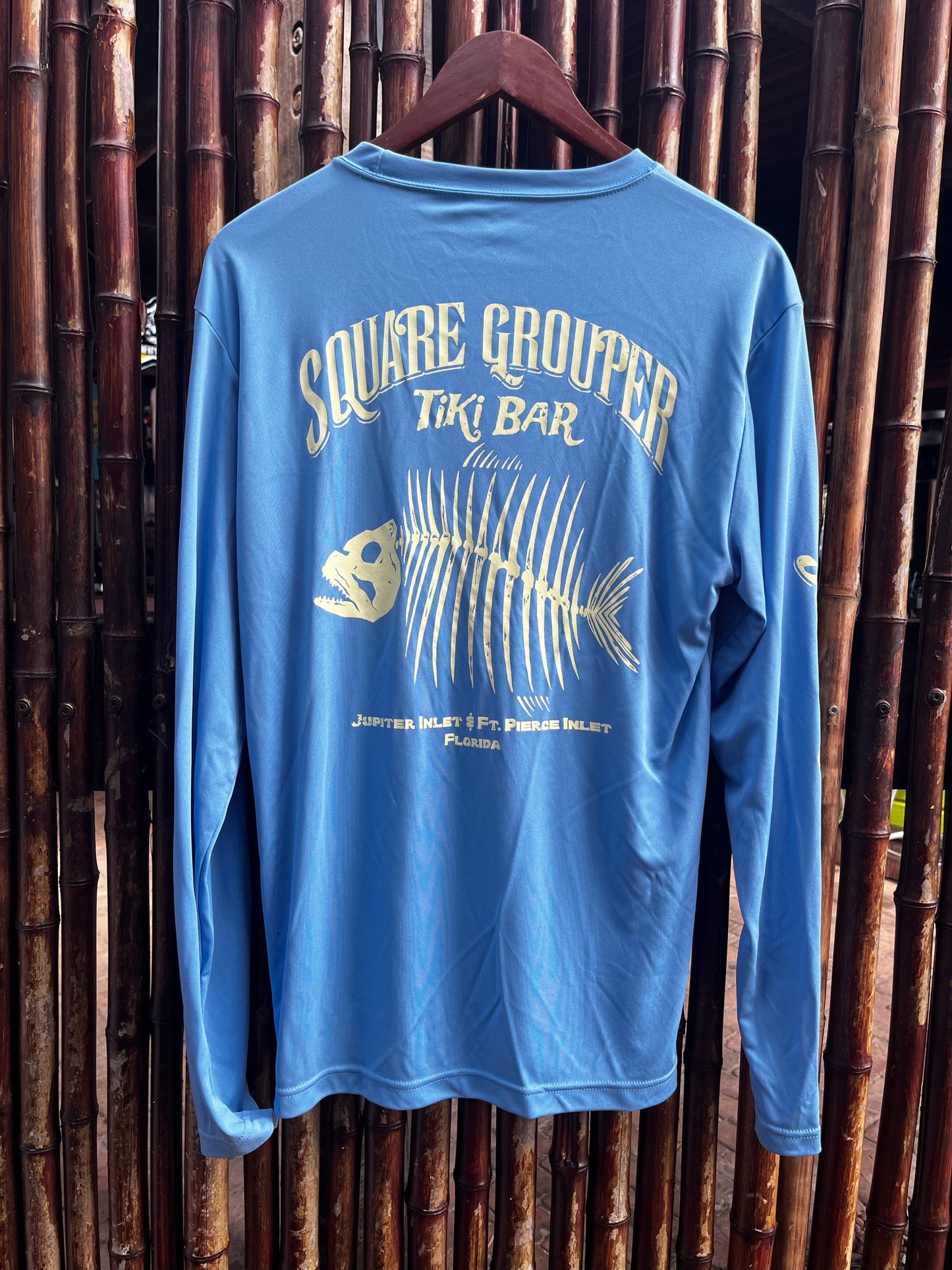 A long sleeve blue tech fabric tee with a white fish skeleton design on the back, Square Grouper logo and sleeves, displayed on a wooden hanger against a bamboo backdrop.