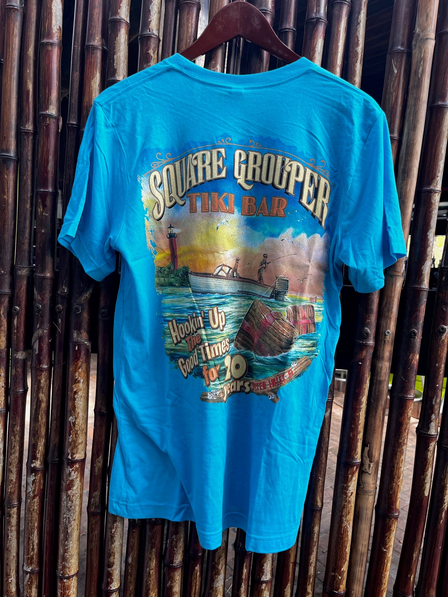 A blue t-shirt with a colorful front print depicting a beach scene and the text 'Square Grouper 20th Year' along with the logo ' Hookin Up The Good Times'.